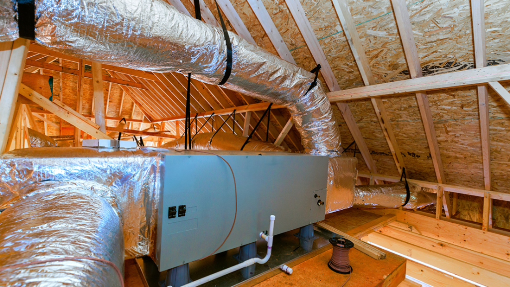 Attic HVAC installation with silver insulated ducts surrounding a gray air handler on a plywood floor, under construction.