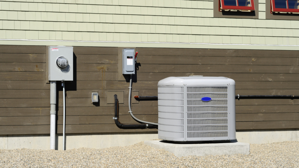 Outdoor air conditioner condenser beside brown wood siding with a meter box and wiring on a gravel yard.