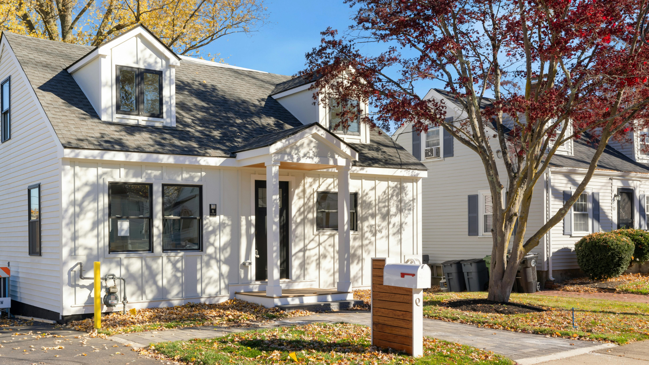 September 2026 | Flader Plumbing White two-story house with front porch, black front door, and fall leaves on the lawn; trees with autumn colors in the background.